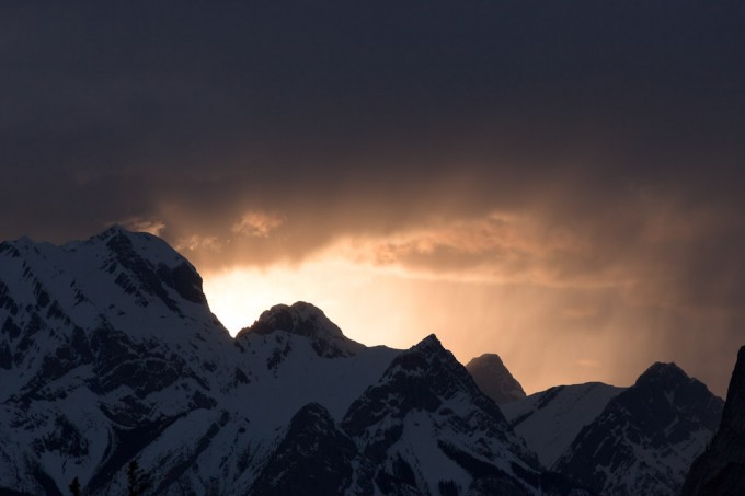 The last light of the sun setting behind rugged peaks shines below a sky of heavy clouds